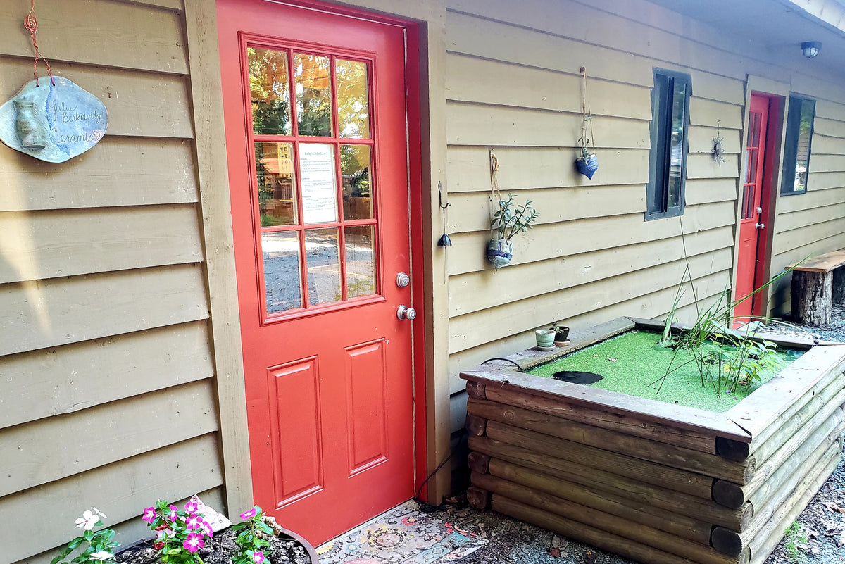The studio’s outside wall: wood siding, the red glass-paned door, the Julie Berkowitz Ceramics sign, hanging planters.