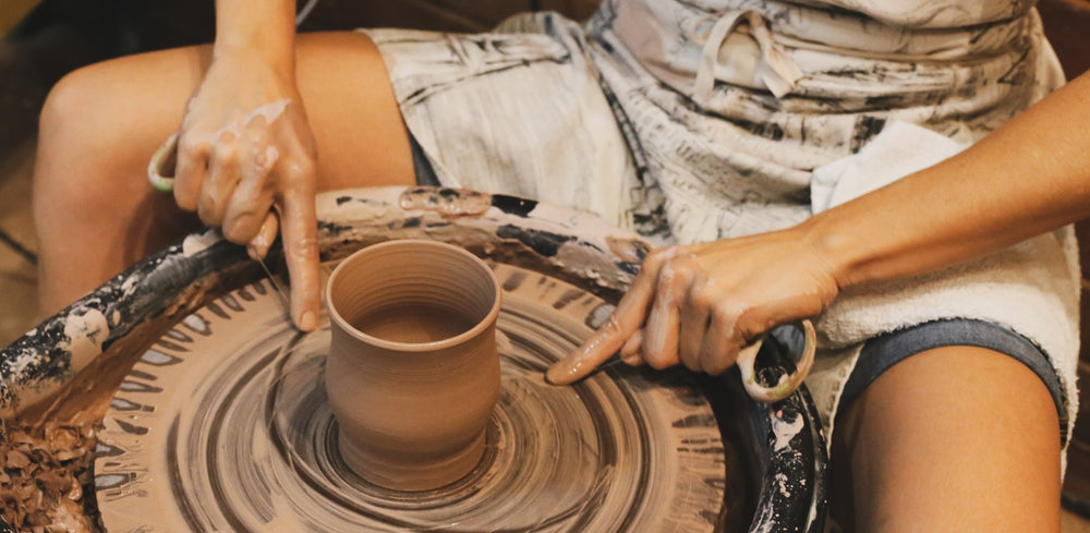 A freshly thrown cup on the wheel, a hand steadying the rim, an apron streaked with clay behind it.