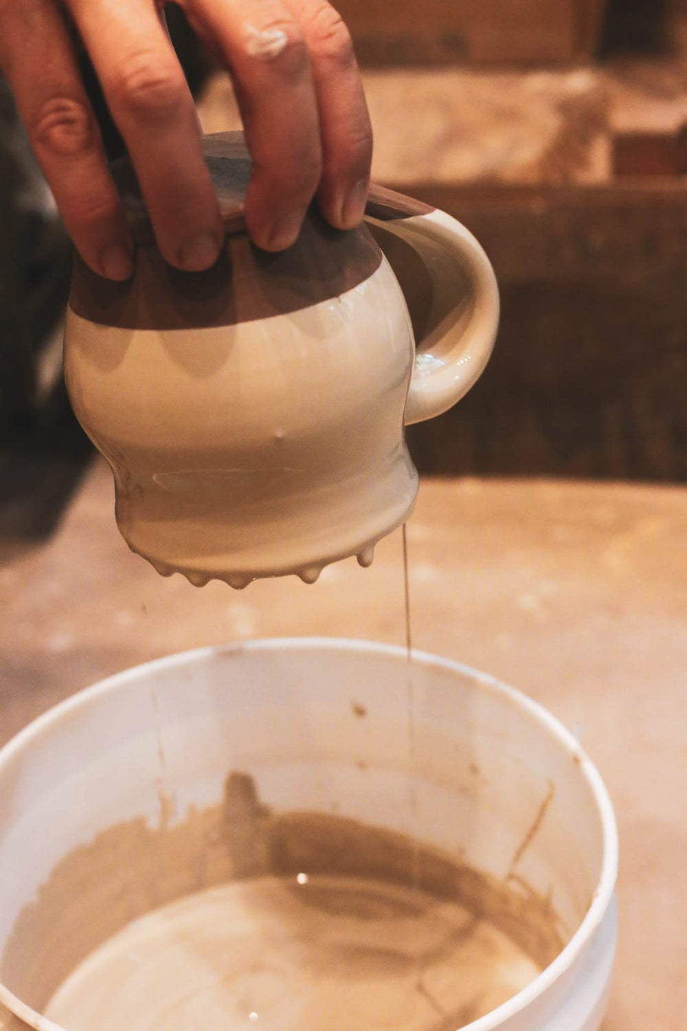 A hand holds a glazed mug upside down over a bucket, glaze dripping from its base.